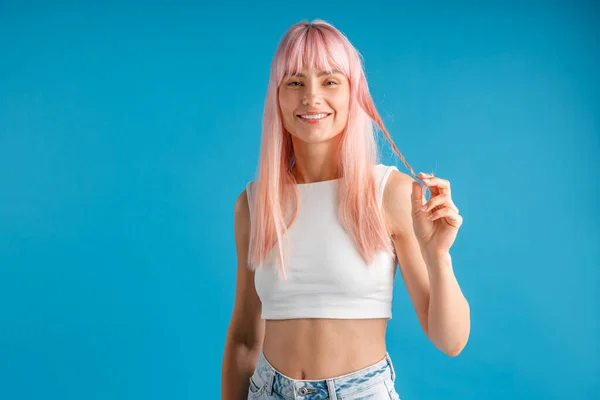 Glimlachende jonge vrouw met natuurlijke lange roze geverfd haar met een streng van het en kijken naar de camera, poseren geïsoleerd over blauwe studio achtergrond — Stockfoto