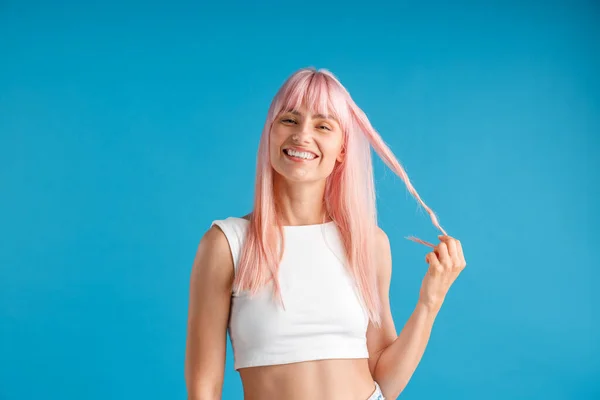 Tevreden jonge vrouw met natuurlijke lange roze geverfd haar met een streng van het en glimlachen op de camera, poseren geïsoleerd over blauwe studio achtergrond — Stockfoto