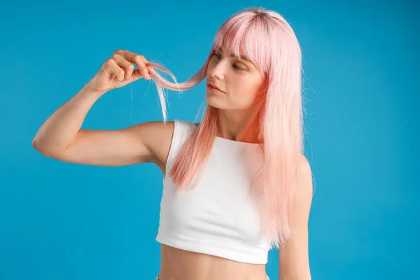 Neugierige Frau, die ihr glattes natürliches langes rosa gefärbtes Haar berührt und ansieht, während sie isoliert vor blauem Studiohintergrund posiert — Stockfoto