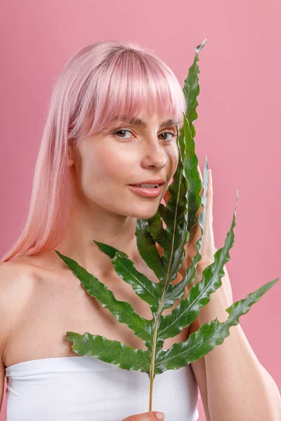Retrato de mujer hermosa con el pelo rosa y la piel perfecta mirando a la cámara, posando con hoja de planta aislada sobre fondo de estudio rosa — Foto de Stock