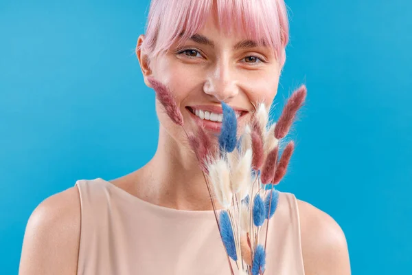 Retrato de una mujer alegre con el pelo rosa sonriendo a la cámara, sosteniendo la Cortaderia seca, hierba de pampas cerca de su cara, posando sobre fondo azul del estudio — Foto de Stock