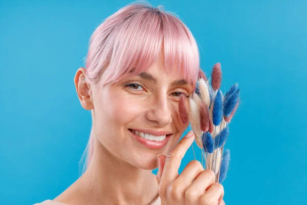 Close-up portret van een mooie vrouw met roze haar glimlachen op de camera, het houden van gedroogde Cortaderia, pampas gras in haar hand, poseren over blauwe studio achtergrond — Stockfoto