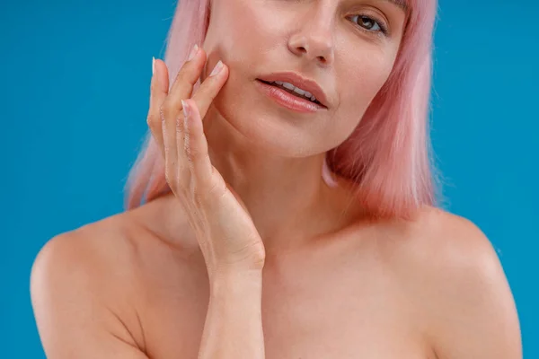 Gros plan de la peau pure et éclatante de la femme avec des cheveux roses touchant son visage, regardant la caméra, posant isolé sur fond bleu studio — Photo