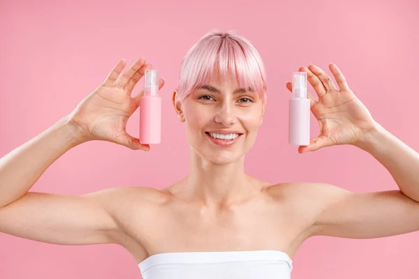 Porträt einer aufgeregten jungen Frau mit rosa Haaren, die lächelnd zwei Plastikflaschen mit Schönheitsprodukten zeigt und isoliert auf rosa Hintergrund posiert — Stockfoto