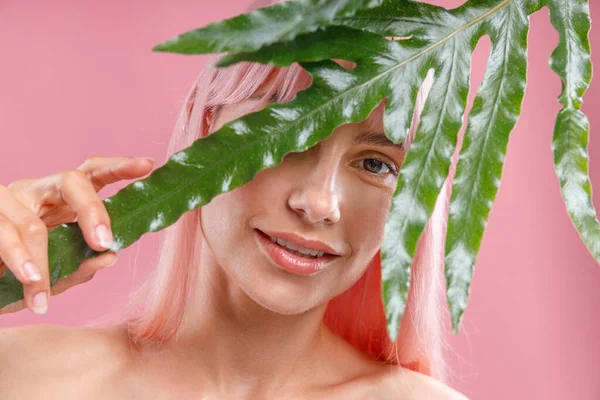 ピンク色の髪の魅力的な女性の肖像画は、カメラを見て、緑の植物の葉からのぞき見、ピンクのスタジオの背景に隔離されたポーズ — ストック写真