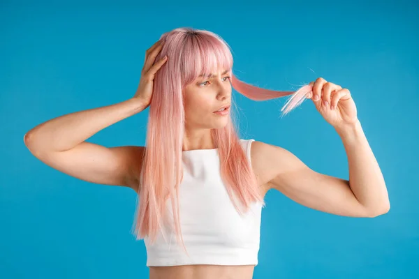 Verwirrte junge Frau, die ihr natürliches langes rosa gefärbtes Haar hält und ansieht, während sie isoliert vor blauem Studiohintergrund posiert — Stockfoto