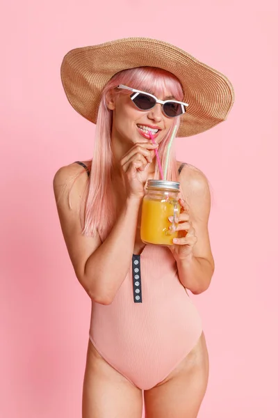 Модная женщина с розовыми волосами в соломенной шляпе, купальнике и солнцезащитных очках пьет коктейль, позируя изолированно на розовом студийном фоне — стоковое фото