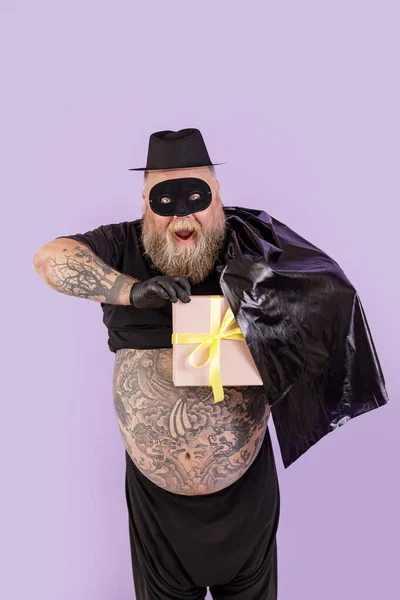 Podekscytowany człowiek z nadwagą w kostiumie Zorro ukrywa pudełko z prezentami za peleryną na fioletowym tle — Zdjęcie stockowe