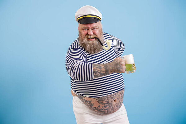 Happy plus size man in sailor suit holds delicious beer on light blue background