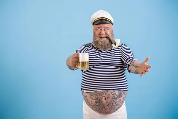 담배 파이프를 든 쾌활하고 뚱뚱 한 선원 이 푸른 배경 위에 신선 한 맥주 한잔을 들고 있다 — 스톡 사진