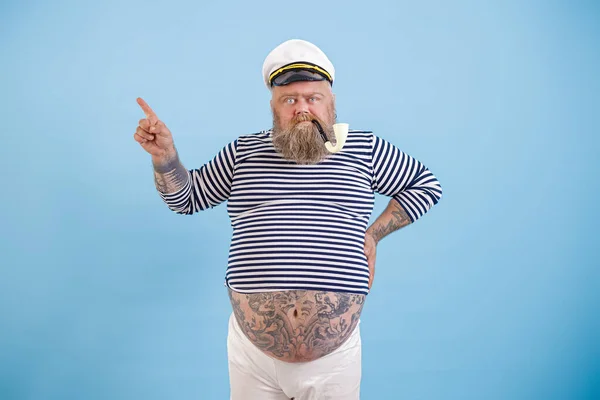 Plus size sailor man with tattoos and smoking pipe points aside by finger on light blue background