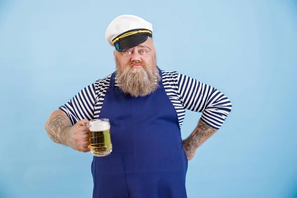 Funny plus size man in sailor suit holds glass of foamy beer on light blue background