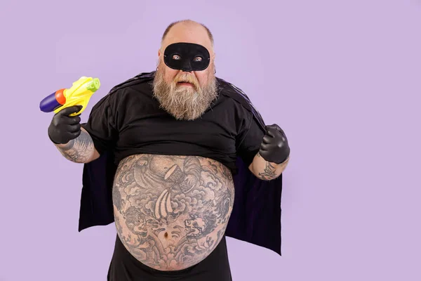 Grappig kwaad zwaarlijvige man in carnaval kostuum houdt speelgoed blaster poseren op paarse achtergrond — Stockfoto