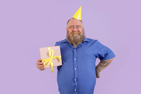 Dojrzały mężczyzna z nadwagą z kapeluszem party posiada pudełko na fioletowe tło — Zdjęcie stockowe