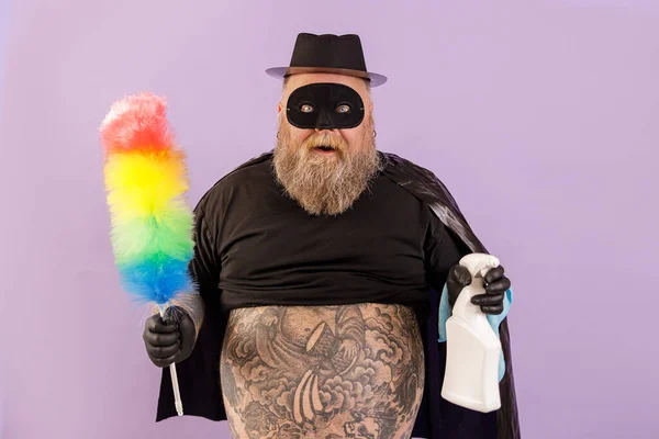Emotionele mollige man in held pak houdt borstel en spray fles op paarse achtergrond — Stockfoto