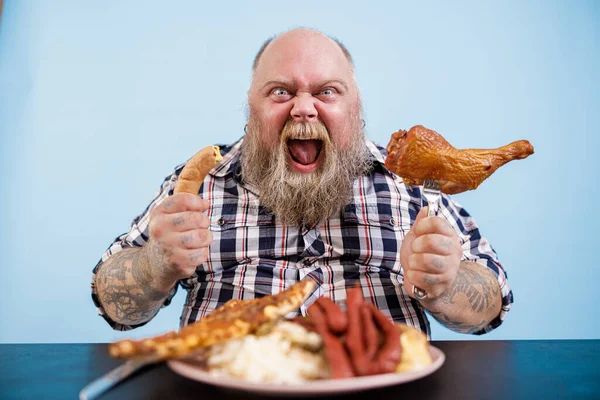 Emotionele zwaarlijvige man houdt worst en gerookte kippenpoot aan tafel — Stockfoto