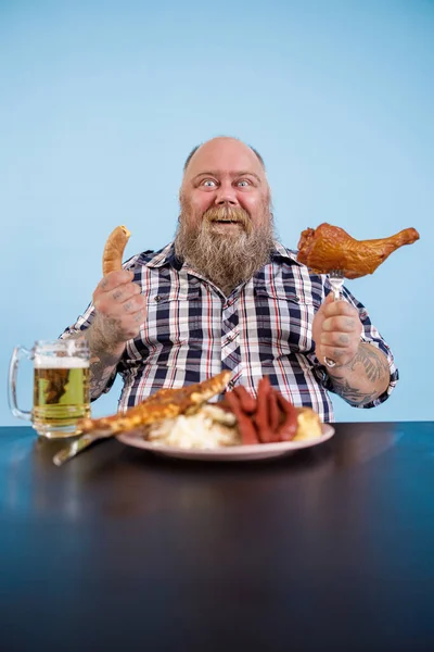 Gut gelaunter Übergewichtiger sitzt mit fettem Essen und Bier am Tisch — Stockfoto