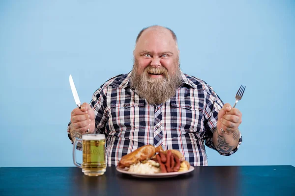 Joyful persoon met overgewicht zit aan tafel met rijk eten en bier op blauwe achtergrond — Stockfoto