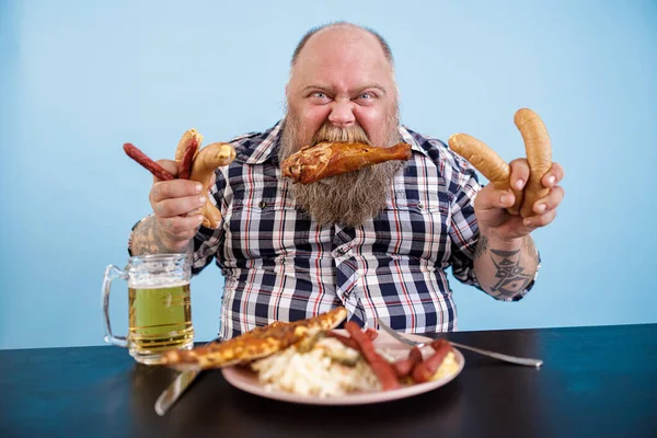 Vette man met kippenpoot in de mond houdt worsten aan tafel in studio — Stockfoto