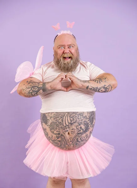 Joyful plus size man in fairy suit shows heart with hands on purple background