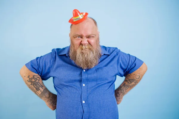 Grappig plus size man draagt kleine sombrero houdt handen op taille grimmig op blauwe achtergrond — Stockfoto