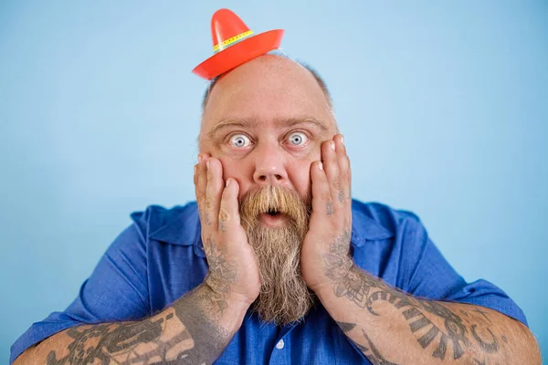 Schockierter fettleibiger Mann mit kleinem Sombrero hält Wangen auf hellblauem Hintergrund — Stockfoto