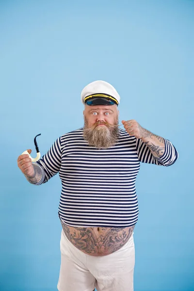 Baard zwaarlijvige man zeeman met roken pijp aanscherpt snor op licht blauwe achtergrond — Stockfoto