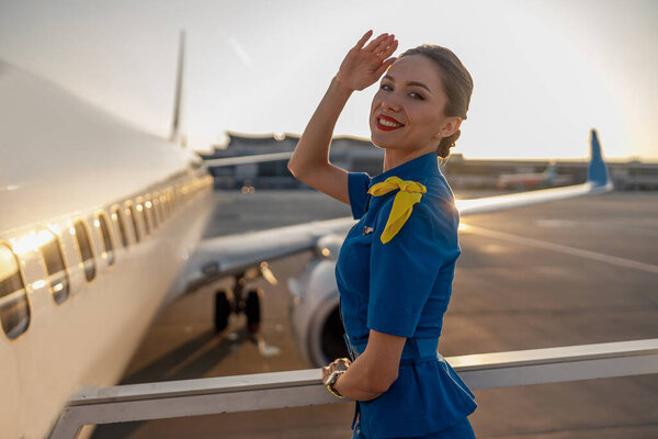 Портрет веселой стюардессы в синей форме, улыбающейся в камеру, позирующей на открытом воздухе на закате. Коммерческий самолет рядом с терминалом в аэропорту на заднем плане