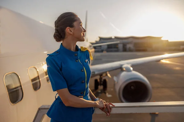 Accurata hostess aerea in uniforme blu che distoglie lo sguardo, in piedi all'aperto al tramonto. Aereo commerciale vicino al terminal in un aeroporto sullo sfondo — Foto Stock