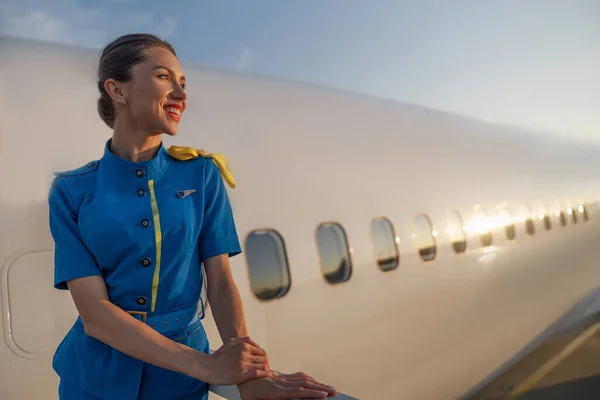 Inspirierte Stewardess in blauer Uniform lächelt zur Seite und steht draußen im Sonnenuntergang. Verkehrsflugzeug im Hintergrund — Stockfoto