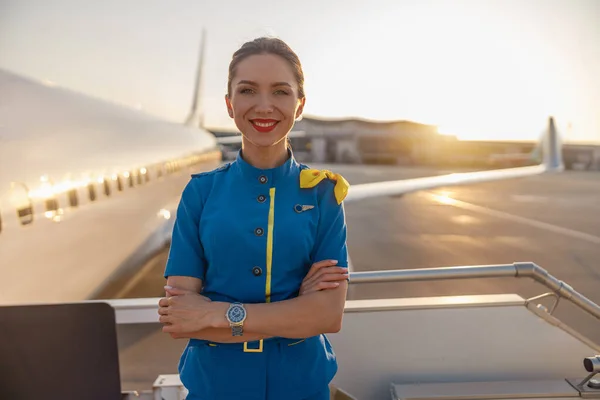 Porträt einer schönen Stewardess mit roten Lippen in blauer Uniform, die in die Kamera lächelt und im Hintergrund mit einem Verkehrsflugzeug in der Nähe des Terminals posiert — Stockfoto