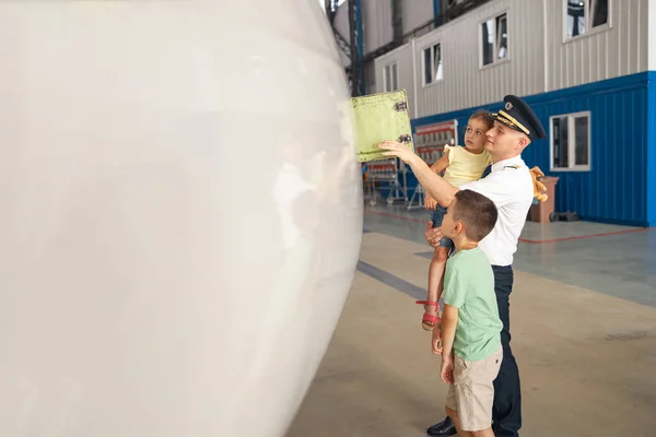 Piloto profesional en uniforme mostrando partes del avión a sus dos niños pequeños que vinieron a visitar a su padre en el hangar de aviones —  Fotos de Stock