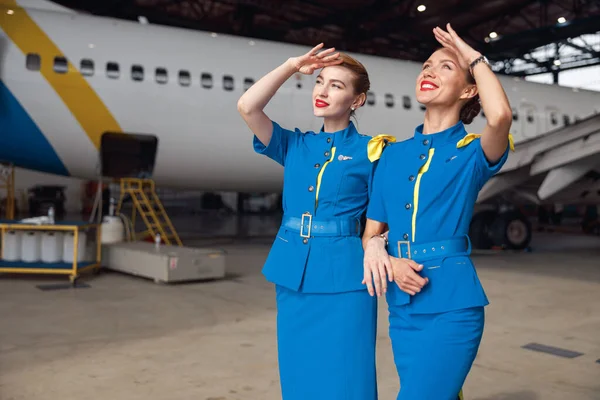 Dvě letušky ve stylové modré uniformě, usmívající se při pohledu na oblohu, stojící společně před osobními letadly v hangáru na letišti — Stock fotografie