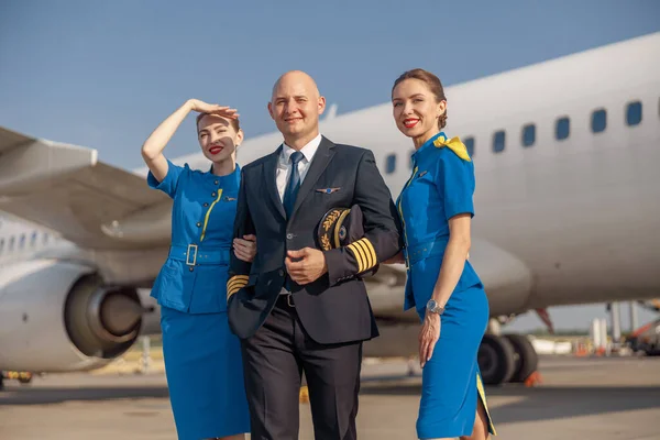 Glücklicher Pilot und zwei attraktive Stewardessen, die zusammen vor einem Flugzeug stehen und nach der Landung lächeln — Stockfoto