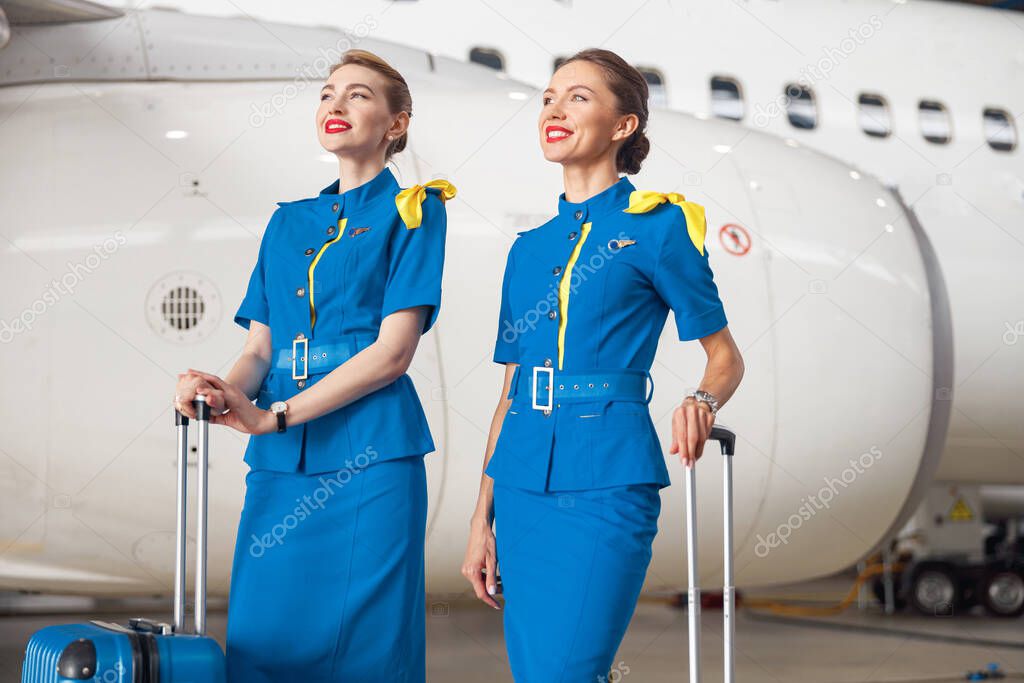 Two confident air stewardesses in bright blue uniform standing with their luggage before departure in front of passenger aircraft in hangar at the airport