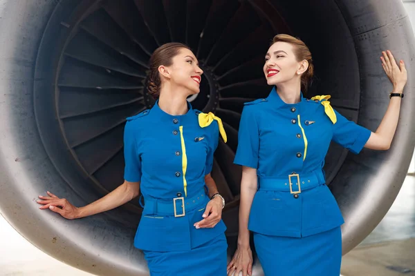 Dvě krásné letušky ve stylové modré uniformě, usmívající se jeden na druhého, stojící společně před letadlovým motorem — Stock fotografie