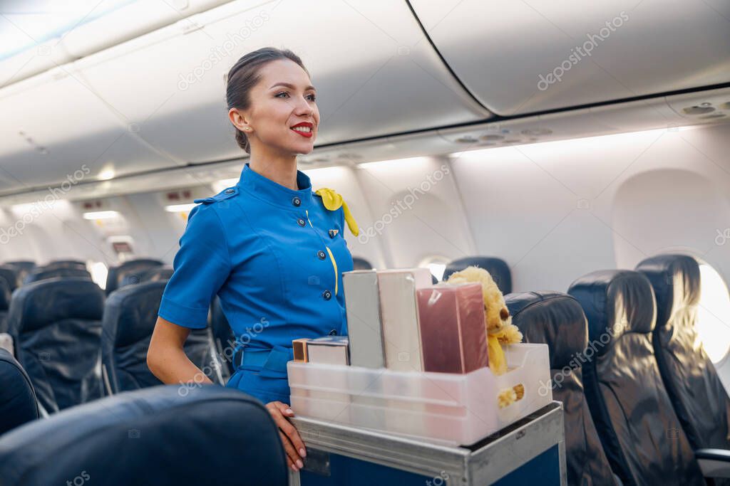 Pretty stewardess in bright blue uniform smiling aside while leading trolley cart with gifts through empty plane aisle