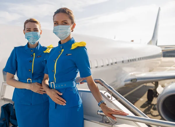 Dvě krásné letušky v modré uniformě a ochranných obličejových maskách při pohledu do kamery, stojící na schodišti při nástupu — Stock fotografie