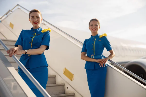 Due belle hostess in uniforme blu che sorridono alla telecamera, in piedi sulla scala aerea e accolgono i passeggeri — Foto Stock