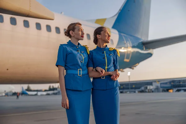 Zwei schöne Stewardessen in blauer Uniform, die bei Sonnenuntergang vor einem großen Passagierflugzeug im Flughafen stehen und weglächeln — Stockfoto