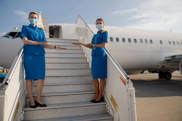 Dlouhý záběr dvou krásných letušek v modré uniformě a ochranných obličejových maskách, jak se dívají do kamery, stojí na schodišti a vítají cestující — Stock fotografie
