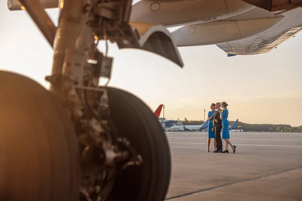 Foto completa de piloto varón posando para sesión de fotos junto con dos azafatas en uniforme azul, de pie en una terminal del aeropuerto al atardecer —  Fotos de Stock