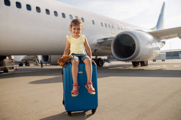 Full length shot of cute little girl holding her toy, looking at camera and sitting on suitcase in front of big airplane