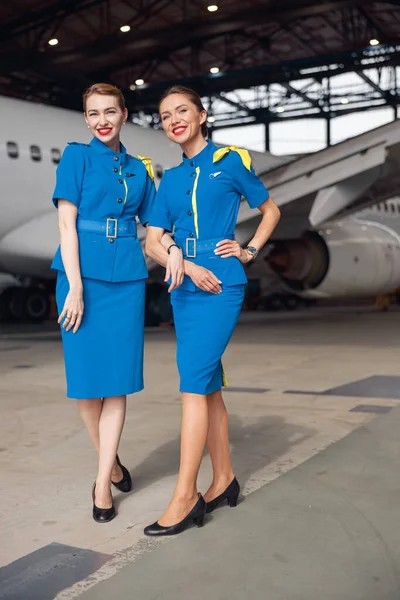 Aufnahme in voller Länge von zwei Stewardessen in stylischer blauer Uniform, die in die Kamera lächeln und gemeinsam vor Passagierflugzeugen im Hangar des Flughafens stehen — Stockfoto