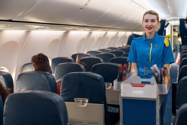 Usmívající se letuška podávající jídlo cestujícím v letadle. Letuška chodící s vozíkem v uličce — Stock fotografie