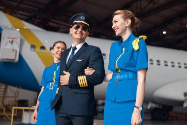 Üniformalı ve havacı güneş gözlüklü gülümseyen pilot havaalanı hangarında mavi üniformalı iki hostesle birlikte büyük yolcu uçağının önünde yürüyor.