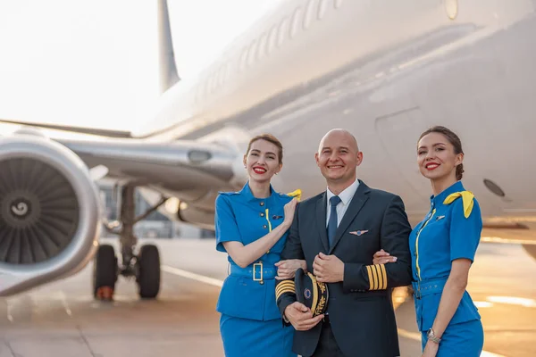 Retrato de un piloto excitado posando junto con dos azafatas en uniforme azul delante de un avión en el aeropuerto —  Fotos de Stock