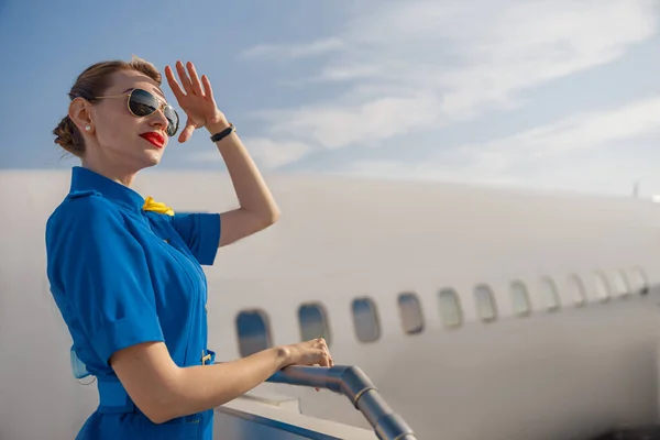Retrato de elegante azafata de aire en uniforme azul y gafas de sol sosteniendo la mano cerca de su cabeza y mirando lejos, de pie en la escalera de aire en un día —  Fotos de Stock