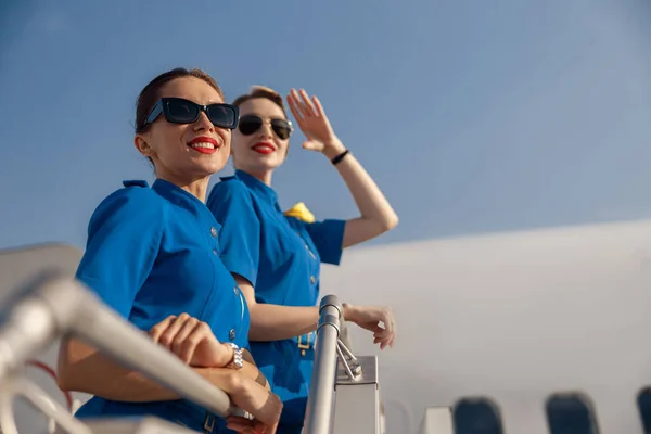 Retrato de dos animadas azafatas en uniforme azul y gafas de sol sonriendo, paradas juntas en la escalera de aire en un día soleado —  Fotos de Stock