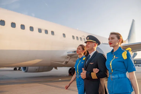 Emocionado piloto masculino caminando junto con dos auxiliares de vuelo en uniforme azul delante de un avión en la terminal al atardecer —  Fotos de Stock